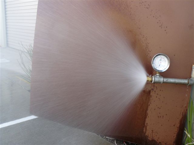 Water spray from a test nozzle fitted with a pressure gauge, discharging against a steel panel
